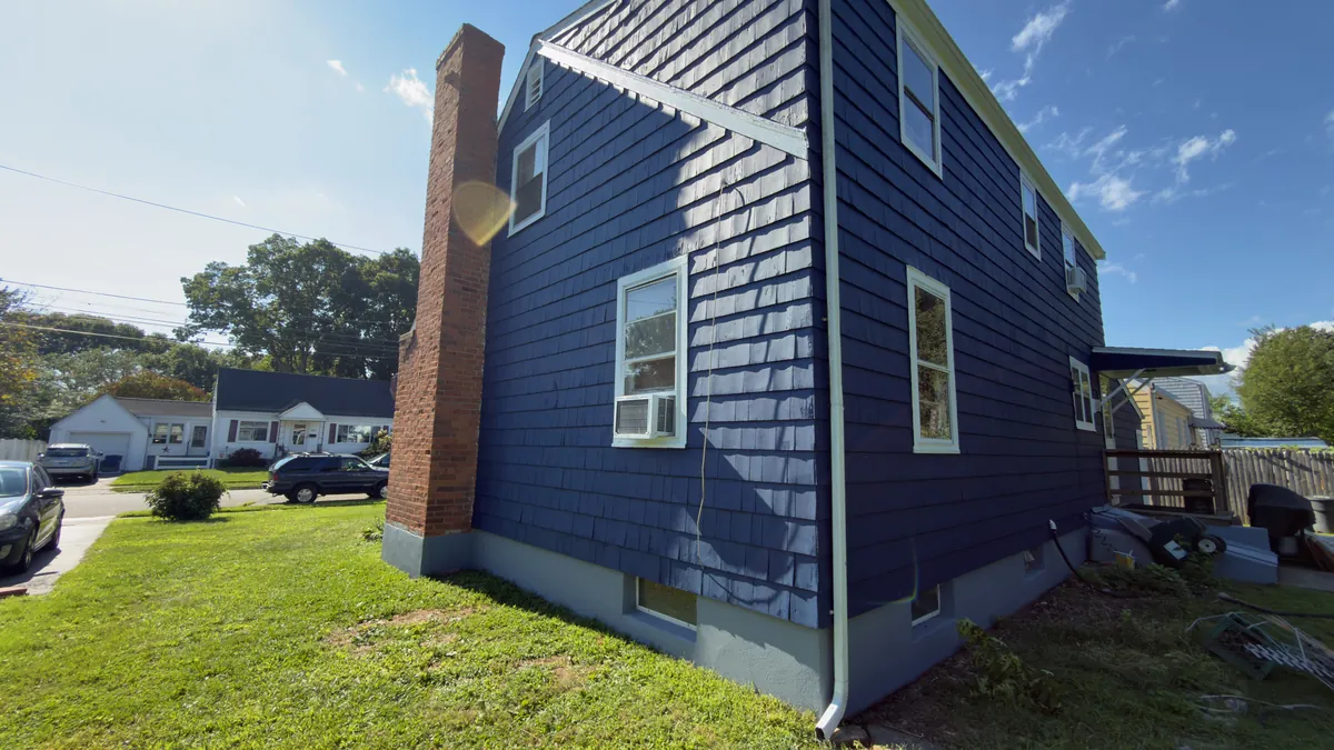 Side of a two-story house with cedar shingle siding painted navy blue, white window trim and a brick chimney in Stratford, CT